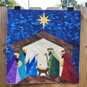 May include: A quilted wall hanging depicting a nativity scene with a bright yellow star against a dark blue starry sky. The scene features figures in purple, teal, blue, green, and red robes gathered around a manger with a baby inside.