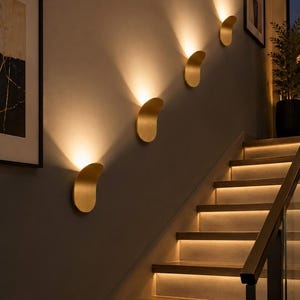 May include: Three curved, gold-toned metal wall sconces are mounted in a diagonal line along a wall. Each fixture emits a warm, upward and downward glow, illuminating the wall surface and the adjacent staircase with integrated step lighting.