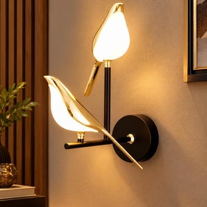 May include: A wall-mounted light fixture featuring two bird-shaped lamps with polished gold-tone bodies and glowing white translucent bellies. The birds are perched on a black metal frame attached to a circular black wall plate, emitting warm light.