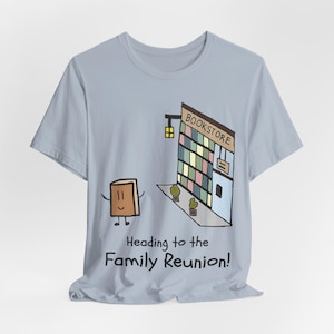 May include: A light blue t-shirt featuring a cartoon illustration of a brown book character walking toward a bookstore building. The text below the graphic reads, "Heading to the Family Reunion!" The shirt has short sleeves and a crew neckline.