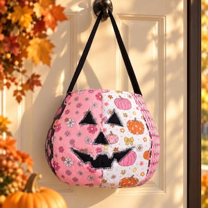 May include: A quilted fabric pumpkin-shaped treat bag with a black handle. The front features a split design: one side is pink with white and pink flowers, the other is white with orange and pink pumpkins, flowers, and bows. It has a black jack-o'-lantern face.