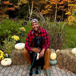May include: A person wearing a red and black buffalo plaid flannel shirt, brown trousers, and a grey baseball cap, crouching while holding a black dog with a white chest patch on a teal leash, surrounded by hay bales and white pumpkins.
