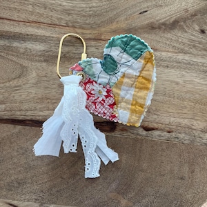 May include: A heart-shaped fabric keychain featuring a patchwork design of yellow gingham, red floral, and green patterned fabric. The heart is attached to a gold-toned metal carabiner and decorated with a white eyelet lace fabric tassel.