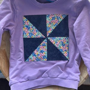 May include: A light purple long-sleeve sweatshirt featuring a square patch on the chest. The patch is divided into eight triangular sections, alternating between dark blue denim fabric and a floral print with pink, blue, and yellow flowers.
