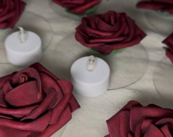 Könnte beinhalten: Eine Ansammlung von künstlichen, dunkelburgunderroten Rosenköpfen, die neben kleinen, weißen, zylindrischen Teelichtern angeordnet sind. Jede Rose weist geschichtete, texturierte Blütenblätter auf, während die Kerzen ein schlichtes Design haben.