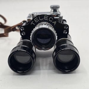 May include: A vintage Teleca opera glass binocular featuring a central chrome lens housing flanked by two black cylindrical barrels. The device includes a black metal bridge with the word TELECA printed in white, a top adjustment knob, and a brown leather strap.