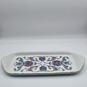 May include: A rectangular white ceramic serving tray with rounded edges and a gold-trimmed rim. The center features a symmetrical floral pattern in shades of blue, red, and green, with intricate leaf and petal motifs arranged in a repeating design.