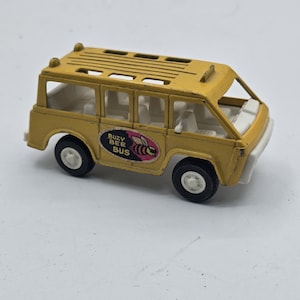 Op de afbeelding: Een gele speelgoedbus met witte interieurstoelen en zwarte wielen. De zijkant heeft een ovale sticker met een bijengrafiek en de tekst BUZY BEE BUS. Het dak heeft een sleufontwerp en de voorbumper en voorruit zijn wit van kleur.