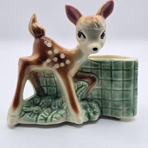 May include: A ceramic deer figurine planter featuring a brown fawn with white spots standing next to a green grid-patterned wall. The fawn has large dark eyes, pink-tinted ears, and is positioned over a base decorated with sculpted green flowers.
