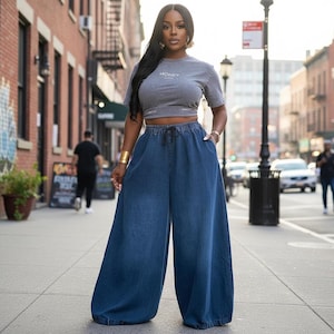Op de afbeelding: Een grijs crop top met korte mouwen en het woord MONEY op de borst, gecombineerd met een donkerblauwe wijde spijkerbroek met een elastische tailleband en een zwart trekkoord. De broek heeft een losse, vloeiende pasvorm en diepe zakken.