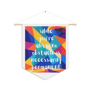May include: A white fabric wall banner hanging from a wooden dowel with a string. The banner features a geometric pattern of multicoloured triangles in red, orange, yellow, blue, and purple, with the white text: while you're alive, no obstacle is necessarily permanent.