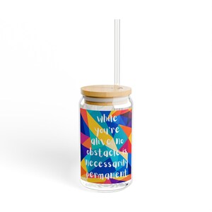 May include: A clear glass tumbler with a bamboo lid and a clear straw. The glass features a vibrant, multi-coloured geometric pattern with the white text: while you're alive, no obstacle is necessarily permanent. The design covers the glass surface.