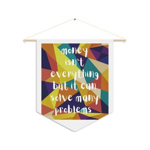 Puede incluir: Un estandarte de pared de tela blanca con una varilla de madera y una cuerda para colgar. El estandarte presenta un patrón geométrico de triángulos en tonos amarillo, naranja, azul y morado, con el texto blanco: money isn't everything but it can solve many problems.