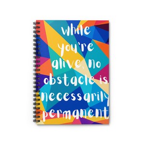 May include: A spiral-bound notebook featuring a vibrant geometric pattern of triangles in shades of blue, yellow, orange, and magenta. The white text printed on the cover reads, "while you're alive, no obstacle is necessarily permanent."