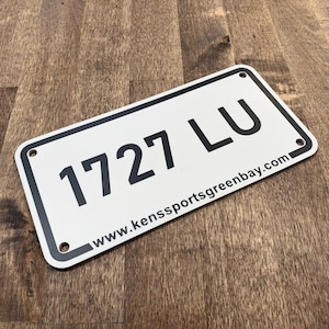 May include: A rectangular white license plate with rounded corners and a black border. The plate features the black text 1727 LU in the center and the website address www.kenssportsgreenbay.com printed in smaller black font along the bottom edge.