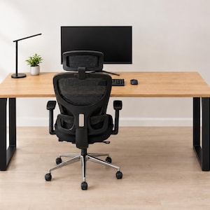 Op de afbeelding: Een rechthoekig bureau van licht hout met zwarte U-vormige metalen poten, gecombineerd met een ergonomische bureaustoel van zwarte mesh met hoofdsteun, verstelbare armleuningen en een vijfpootbasis met wielen, monitor, lamp en plant.