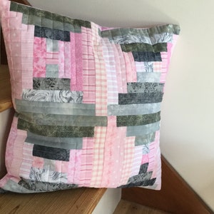 May include: A pink and gray patchwork pillow with a square shape. The pillow is made of different fabrics with various patterns, including stripes, plaid, and floral designs.