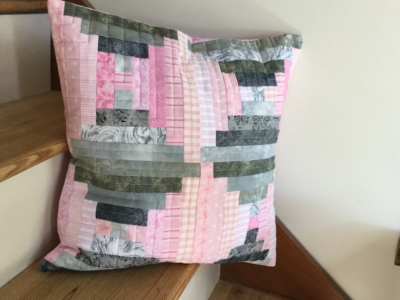 May include: A pink and gray patchwork pillow with a square shape. The pillow is made of different fabrics with various patterns, including stripes, plaid, and floral designs.