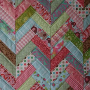 May include: A patchwork quilt with a chevron pattern in shades of pink, green, blue, and brown. The quilt is made of various fabrics with different patterns, including polka dots, stripes, and floral prints.