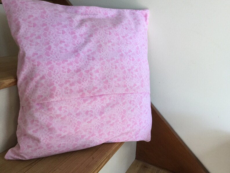 May include: A pink pillow with a white heart pattern. The pillow is on a wooden surface.