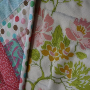 May include: A white quilt with a floral pattern in shades of pink, yellow, and green. The quilt is made of fabric squares sewn together to create a patchwork design.