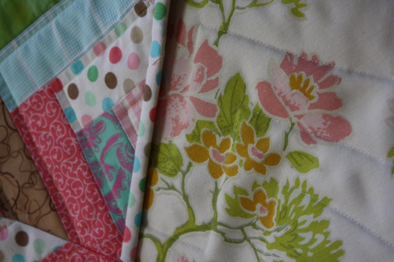 May include: A white quilt with a floral pattern in shades of pink, yellow, and green. The quilt is made of fabric squares sewn together to create a patchwork design.