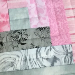 May include: A close-up of a quilt made with pink and gray fabric. The quilt is made of squares and rectangles of different sizes and patterns. The fabric is a mix of solid colors, floral prints, and stripes.
