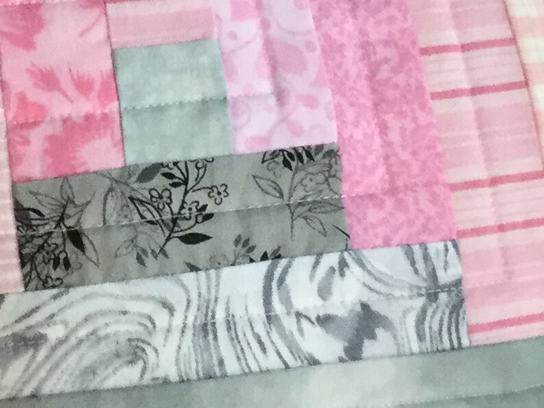May include: A close-up of a quilt made with pink and gray fabric. The quilt is made of squares and rectangles of different sizes and patterns. The fabric is a mix of solid colors, floral prints, and stripes.