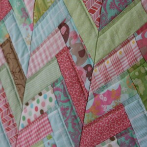 May include: A patchwork quilt with a chevron pattern in shades of pink, green, and blue. The quilt is made of various fabrics with different patterns, including polka dots, gingham, and floral prints.