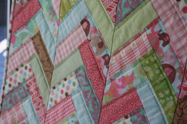 May include: A patchwork quilt with a chevron pattern in shades of pink, green, and blue. The quilt is made of various fabrics with different patterns, including polka dots, gingham, and floral prints.