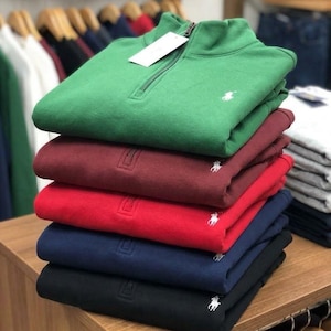 Peut inclure: Une pile de cinq pulls à fermeture éclair au col, déclinés en vert, bordeaux, rouge, bleu marine et noir. Chaque pull présente un petit logo blanc brodé sur la poitrine gauche et une fermeture éclair en métal. Une étiquette blanche est fixée.