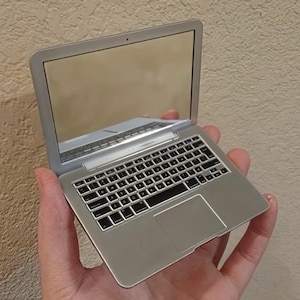 Puede incluir: Un pequeño espejo compacto con forma de ordenador portátil plateado, con una superficie de pantalla reflectante, un teclado negro y un panel táctil liso, diseñado para imitar la apariencia de un dispositivo informático portátil.