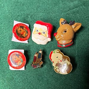 May include: A collection of six holiday pins including two round red pins with the text Keep Christ in Christmas, a Santa Claus face pin, a brown reindeer head pin with a red nose, a candle pin, and a brown bear pin wearing a Santa hat.