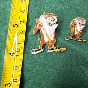 May include: Two enamel pins featuring the Tasmanian Devil character holding a yellow hockey stick and wearing black sneakers. A yellow measuring tape is positioned to the left, showing the larger pin is approximately 1.75 inches in height.