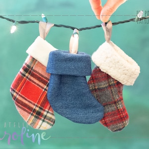 Op de afbeelding: Drie kerstsokken hangen aan een lichtsnoer. Eén sok is rood-zwart geruit, één is van blauwe denim en één is rood-grijs geruit met een witte nep bontkraag. De tekst "PDF PATTERN PATRON PDF" is zichtbaar in de rechterbovenhoek van de afbeelding.