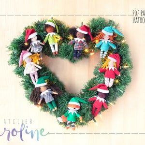 May include: A Christmas wreath made of green garland with a heart shape in the center.  The wreath is decorated with nine handmade felt elf dolls wearing red, green, and blue outfits.  The dolls have different hair colors and skin tones.  The wreath is decorated with white fairy lights.  The text "PDF PATTERN PATRON PDF" is in the upper right corner of the image.  The text "ATELIER Caroline" is in the lower left corner of the image.