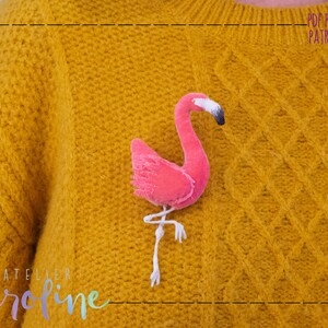 May include: A pink flamingo brooch with white legs and a black beak, pinned to a yellow knitted sweater. The sweater has a cable knit pattern and the text "Atelier Caroline" is visible at the bottom.