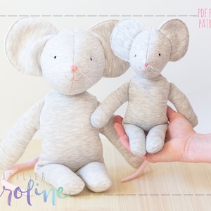 May include: Two grey fabric mouse plushies, one larger than the other, with pink noses and embroidered whiskers. The larger mouse is sitting on a wooden surface with its tail curled behind it. The smaller mouse is being held by a hand.