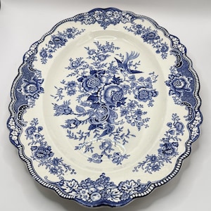 May include: An oval ceramic platter featuring a white base with a blue transferware pattern. The design includes intricate floral arrangements, scrolling foliage, and a central bird motif. The scalloped rim is decorated with a repeating floral border.