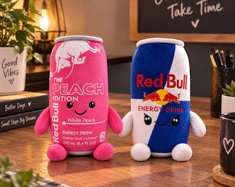 May include: Two plush toys shaped like energy drink cans. The left one is pink with white text reading Red Bull The Peach Edition White Peach 250 mL 8.4 FL OZ. The right one is blue and white with red text reading Red Bull Energy Drink.