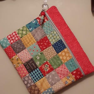 May include: A rectangular fabric pouch featuring a patchwork design of various colorful squares with floral, plaid, and text patterns. It has a red textured border, a gray zipper, and a silver metal ring pull with red, white, and gray beads.