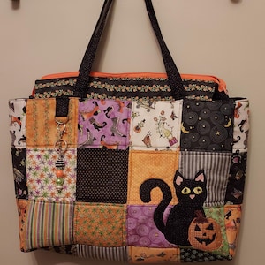 Può includere: Una borsa tote trapuntata con design patchwork a tema Halloween nei colori arancione, nero e viola. La borsa presenta un'applicazione di un gatto nero che tiene una zucca e un ciondolo decorativo di perline su un anello metallico frontale.