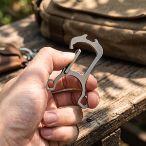 May include: A matte grey metal multi-tool carabiner featuring a spring-loaded gate, a bottle opener notch at the top, and two smaller hook-shaped extensions at the bottom. The tool has a smooth, industrial finish and a geometric, angular frame.