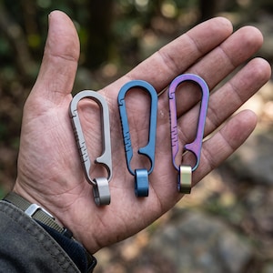 May include: Three metal carabiner clips with a spring-loaded gate mechanism and a small attached loop. The carabiners are finished in matte grey, solid blue, and a multi-colored iridescent gradient, each featuring textured grip notches on the side.