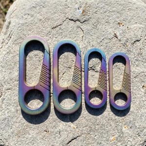 Könnte beinhalten: Vier ovale Karabiner mit einem irisierenden Farbverlauf von Blau über Violett bis Gold. Jeder Karabiner verfügt über einen federbelasteten Verschlussmechanismus und eine kreisförmige untere Öse, angeordnet in absteigender Größe von links nach rechts.