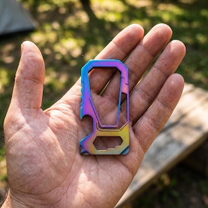 May include: A multi-tool carabiner featuring an iridescent, rainbow-colored metal finish. The tool includes a spring-loaded gate, a bottle opener cutout on the side, and a hexagonal opening at the base, all integrated into a single flat frame.