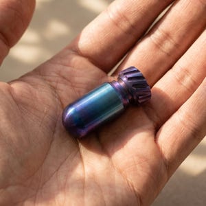 May include: A small, cylindrical metal object with a smooth, iridescent blue and purple finish. The item features a rounded end and a textured, gear-like cap at the opposite side, showcasing a metallic gradient color transition.