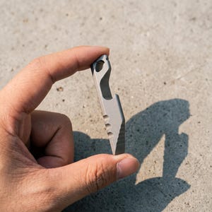 May include: A small, matte grey metal multi-tool featuring a bottle opener cutout at the top, a circular hole for a lanyard, and three distinct semi-circular indentations along the side edge, ending in a tapered, flat-edged tip.