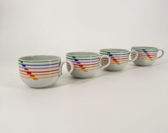 May include: Four white ceramic cups arranged in a row, each featuring a series of horizontal stripes in blue, purple, red, and orange, intersected by a single diagonal orange stripe on the side, with a curved handle on the right side.