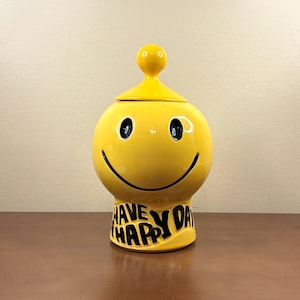 May include: A bright yellow ceramic jar with a lid featuring a round knob. The jar has a black smiley face design and the black text HAVE A HAPPY DAY printed on the base. The container has a rounded body and a wide, stable bottom section.